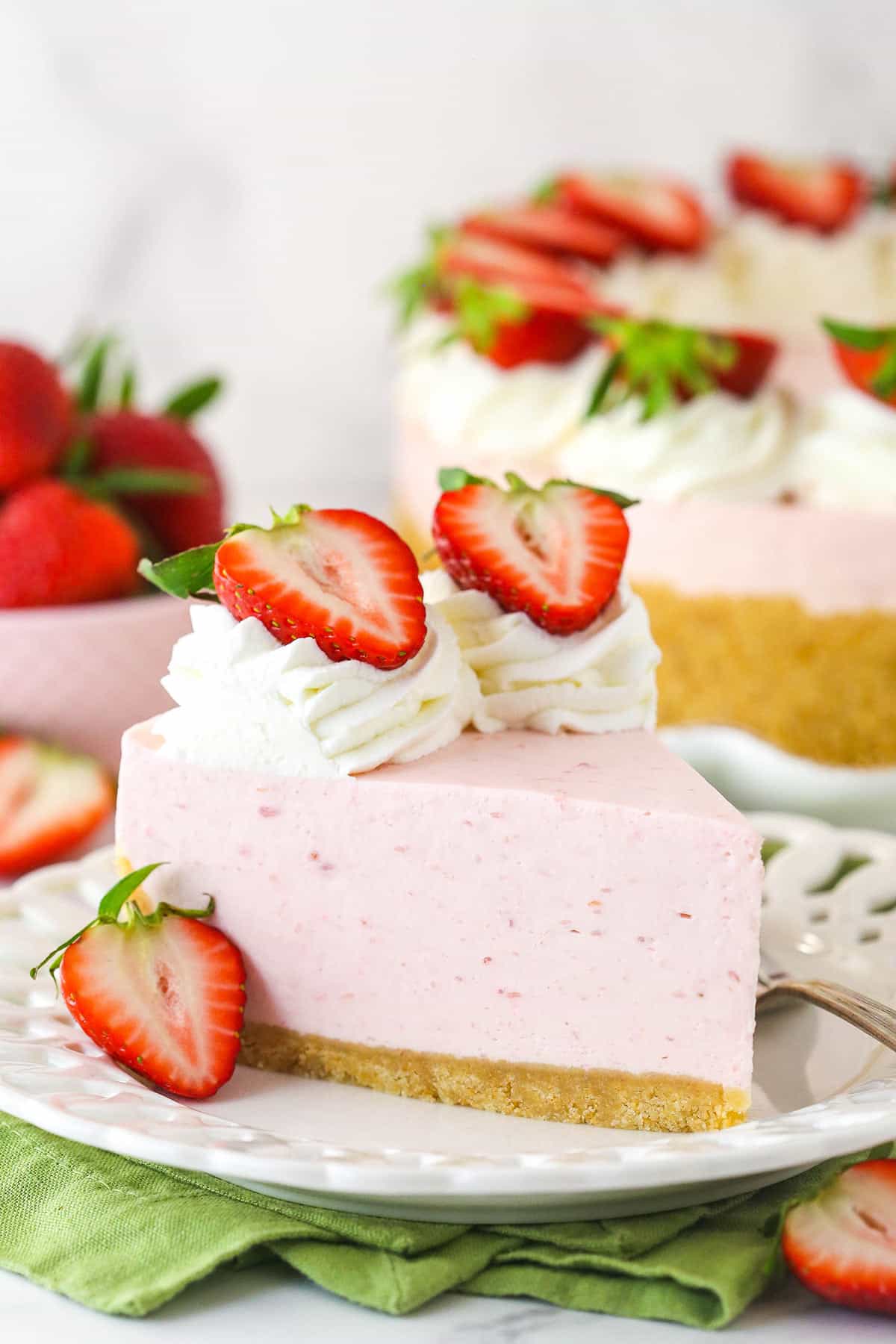 A slice of no bake strawberry cheesecake served on a plate with a fork near a full cheesecake and a bowl of fresh strawberries.