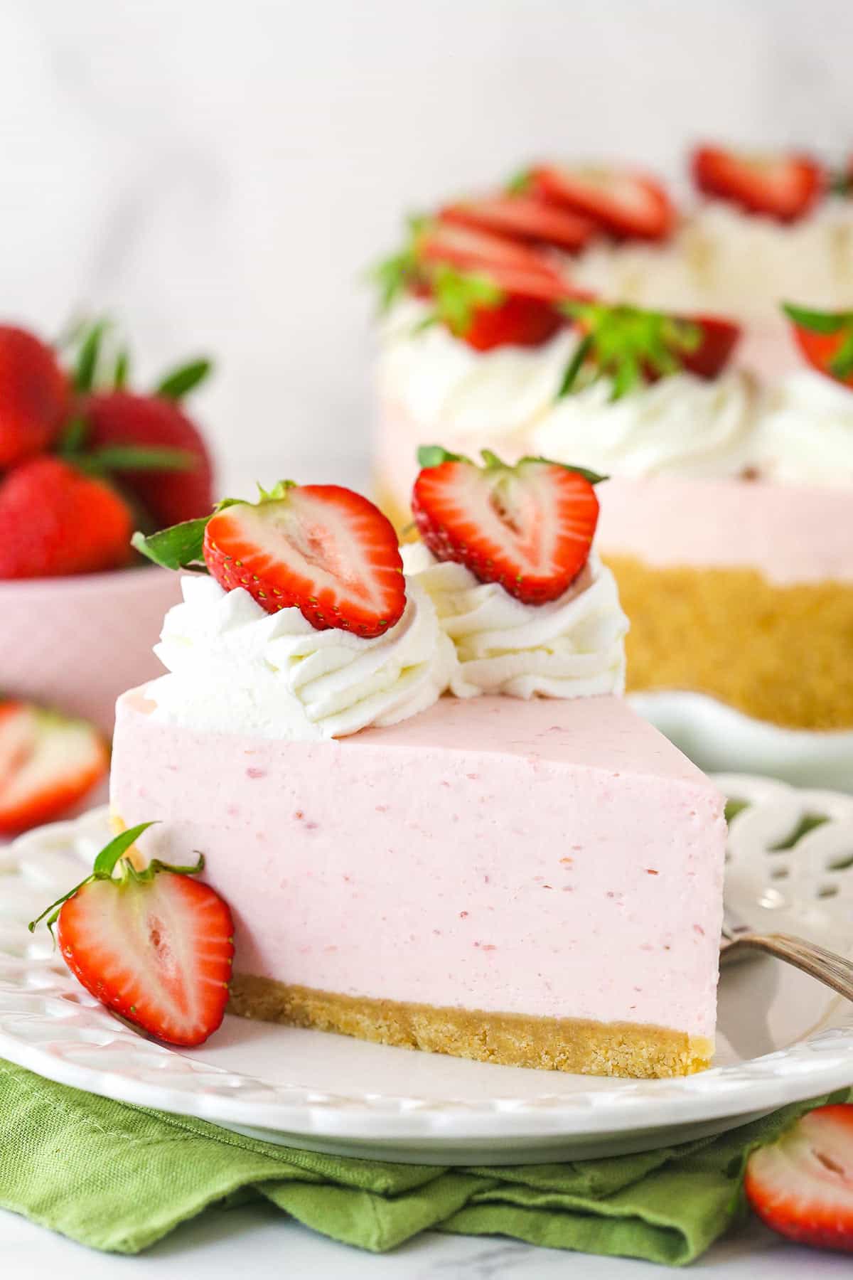 This no bake strawberry cheesecake is made of buttery vanilla wafer crust, creamy strawberry filling, and homemade whipped cream. A slice of no bake strawberry cheesecake served on a plate with a fork near a full cheesecake and a bowl of fresh strawberries.