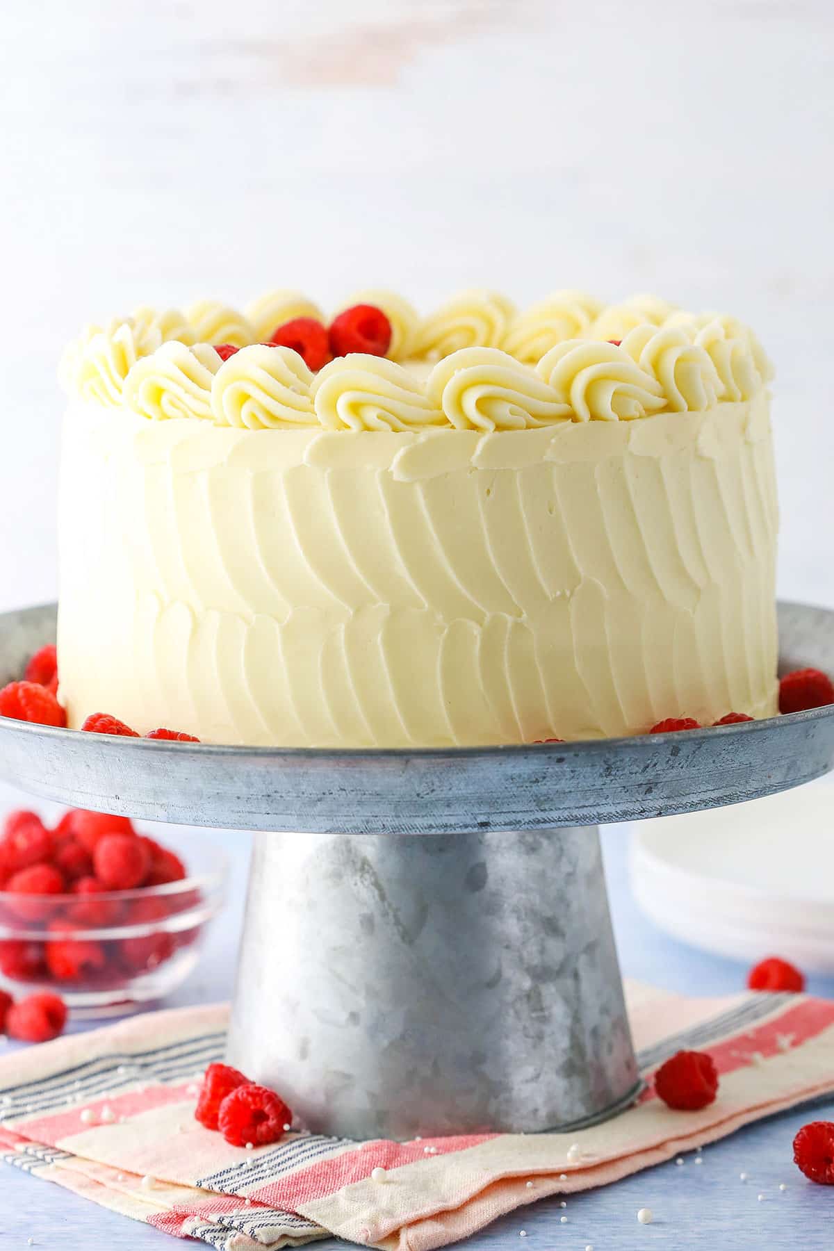 Side view of a full Raspberry Dream Cake on a silver cake stand surrounded by raspberries