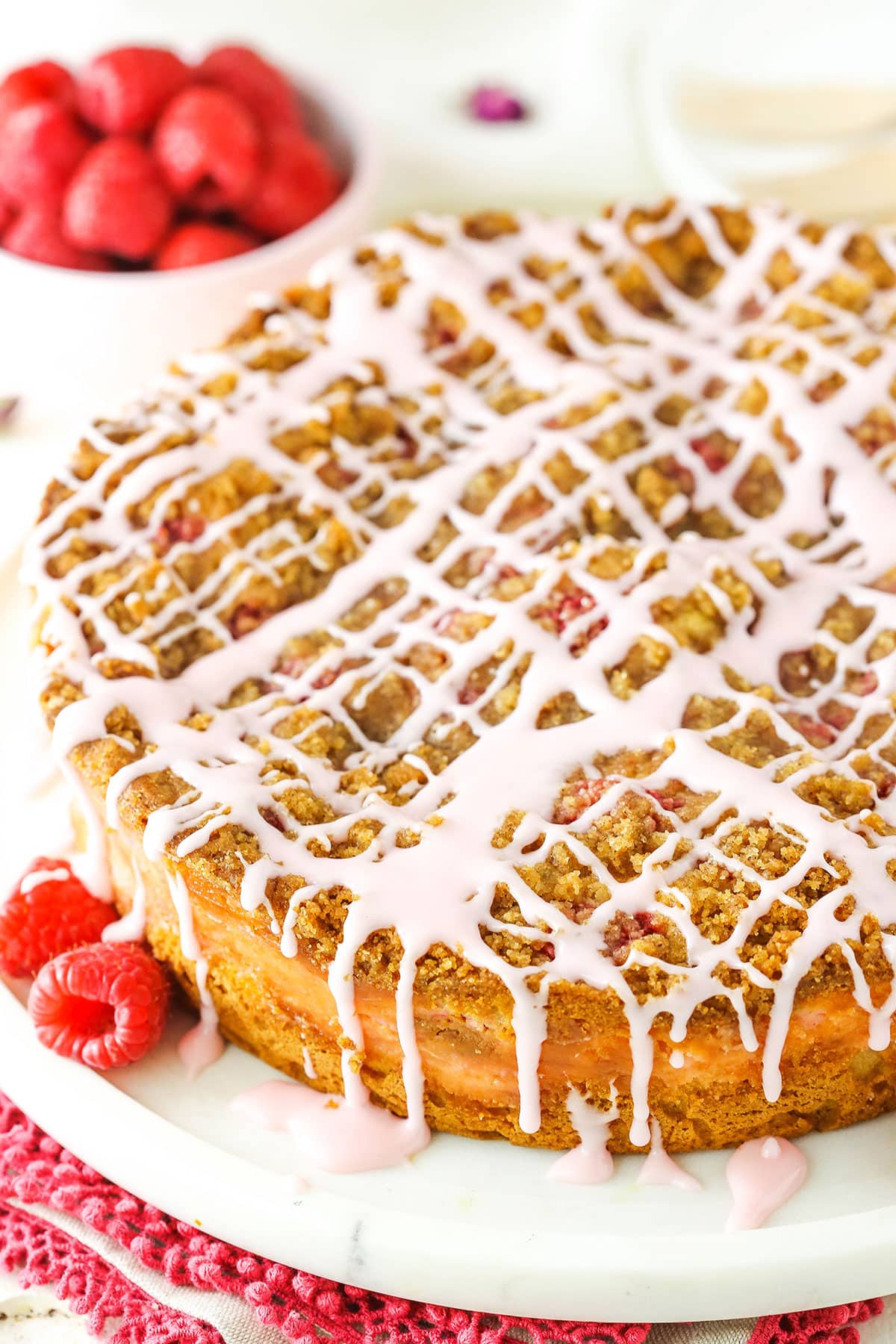 This Raspberry Rose Coffee Cake recipe is such an amazing flavor combination! The coffee cake is filled with a vanilla rose cream cheese filling and fresh raspberries, then topped with cinnamon streusel and a rose glaze! A Raspberry Rose Coffee Cake on a white serving platter with raspberries in the background