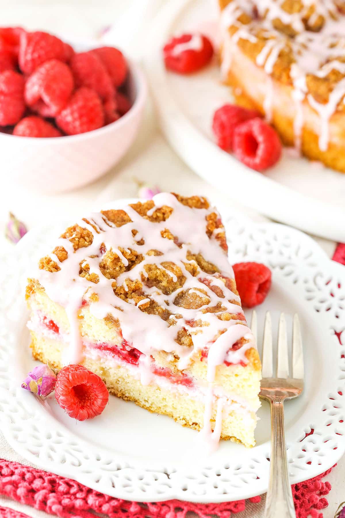 A slice of Raspberry Rose Coffee Cake next to a fork on a white plate with raspberries in the background