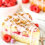 A slice of Raspberry Rose Coffee Cake next to a fork on a white plate with raspberries in the background