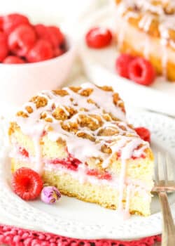 A slice of Raspberry Rose Coffee Cake next to a fork on a white plate with raspberries in the background