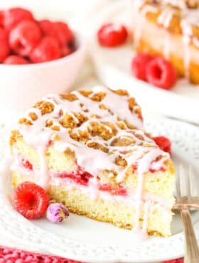 A slice of Raspberry Rose Coffee Cake next to a fork on a white plate with raspberries in the background