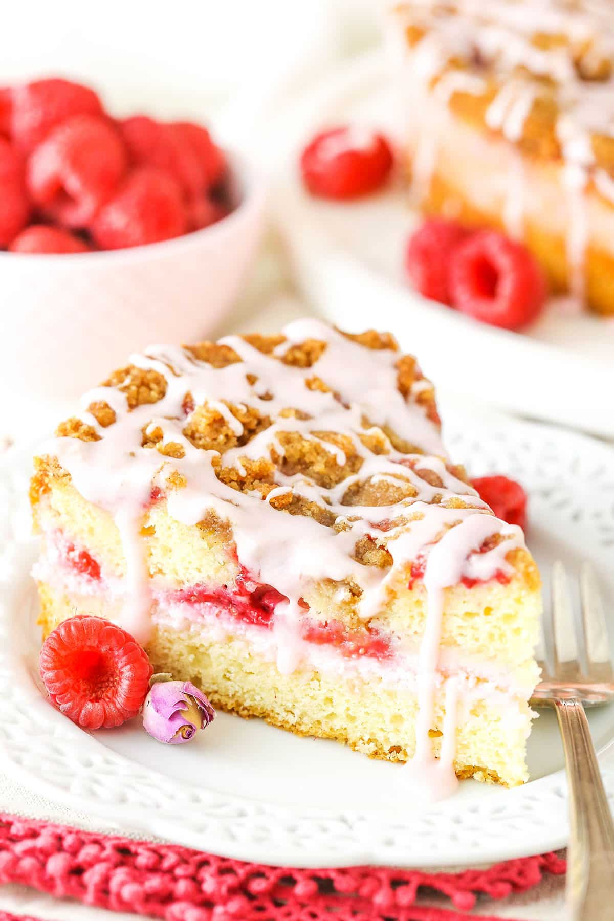 A slice of Raspberry Rose Coffee Cake next to a fork on a white plate with raspberries in the background