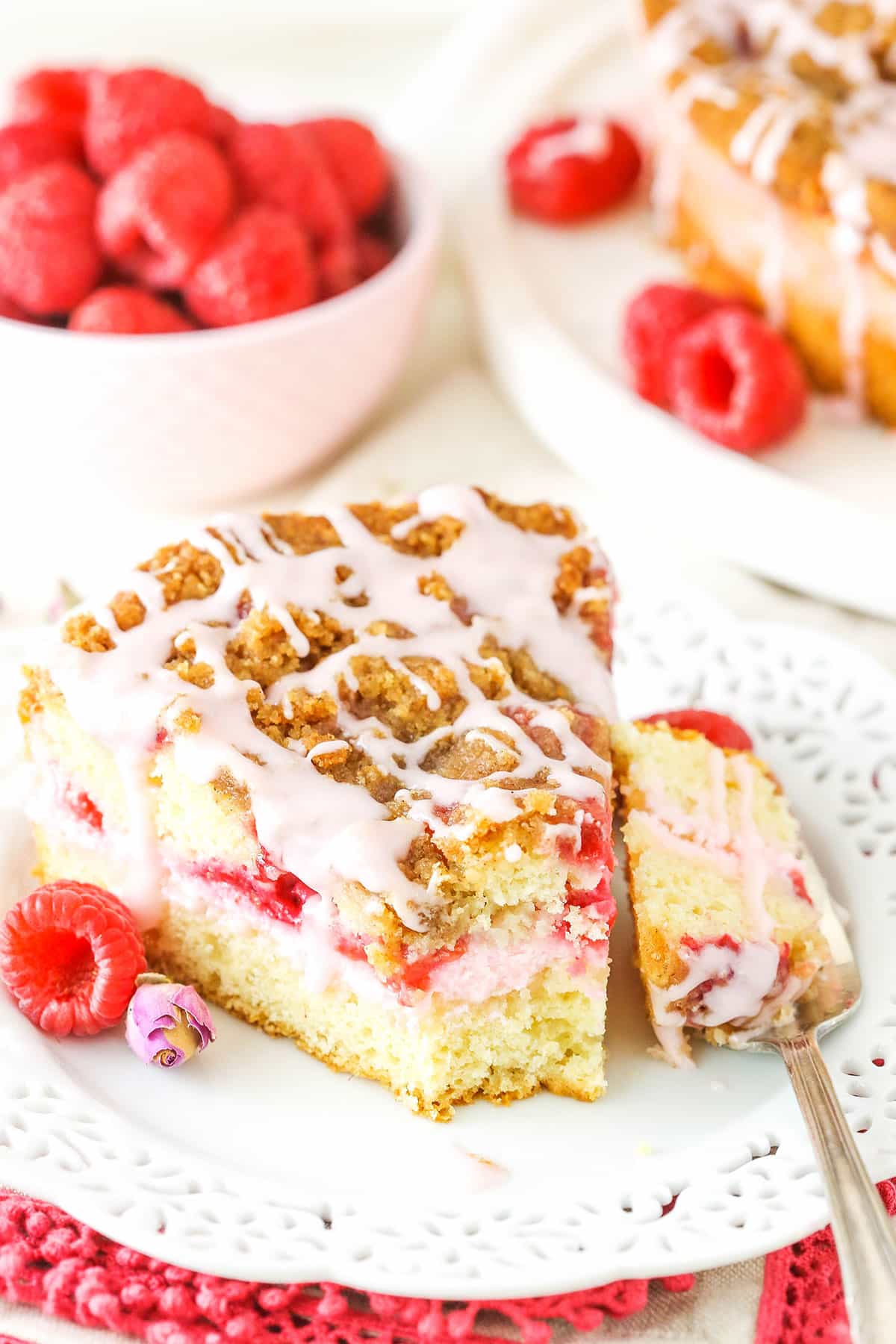 A slice of Raspberry Rose Coffee Cake with a bite taken out next to a fork on a white plate with raspberries in the background