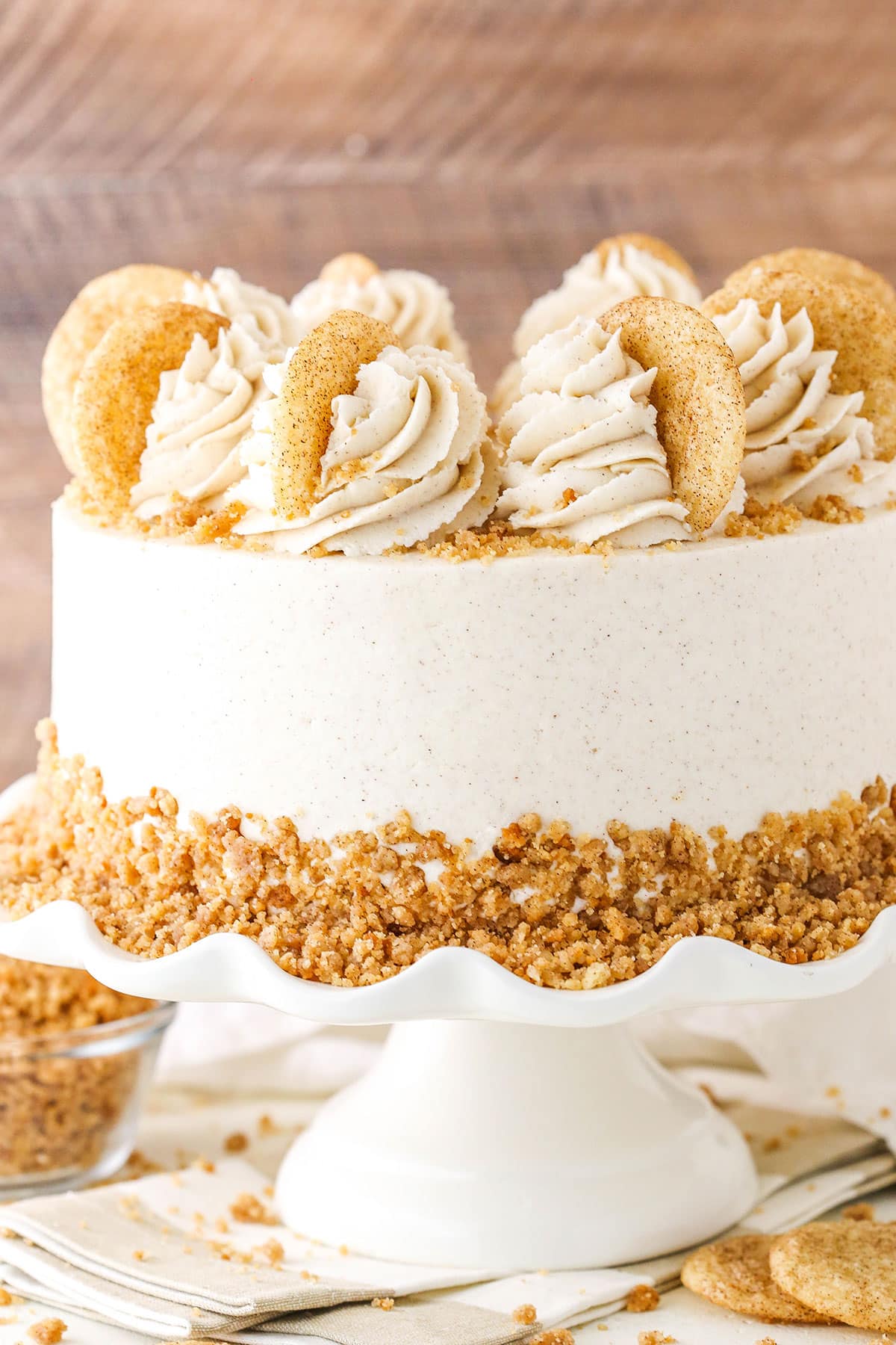 This easy Snickerdoodle Layer Cake is full of cinnamon and sugar! Four layers of moist cinnamon cake are layered with a snickerdoodle crumble and snickerdoodle cookie filling. It’s all covered with cinnamon buttercream for one amazing cake that would be an amazing Thanksgiving or Christmas dessert! Side view of a full Snickerdoodle Layer Cake on a white cake stand