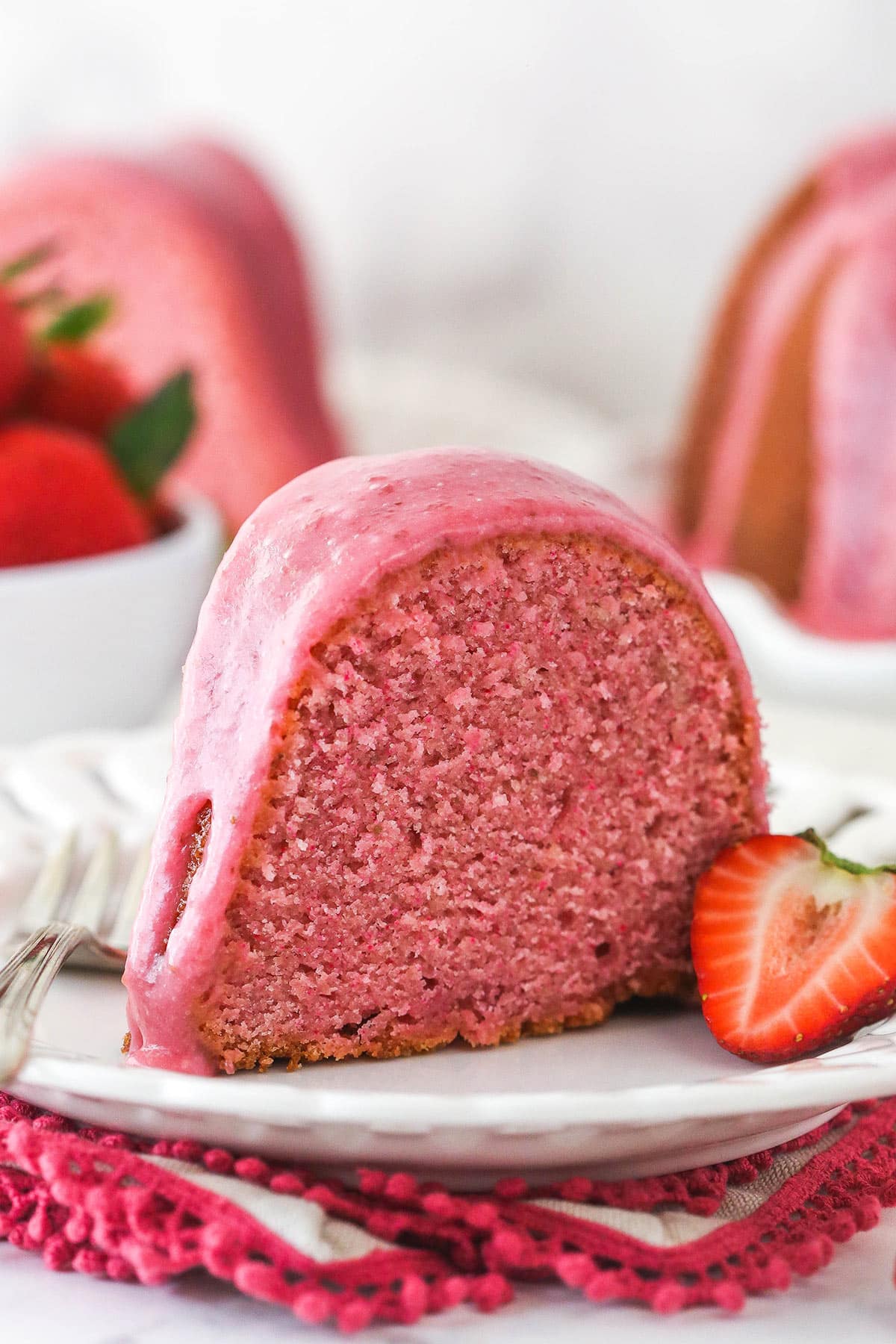 A slice of strawberry pound cake on a plate with a fork and half a fresh strawberry.