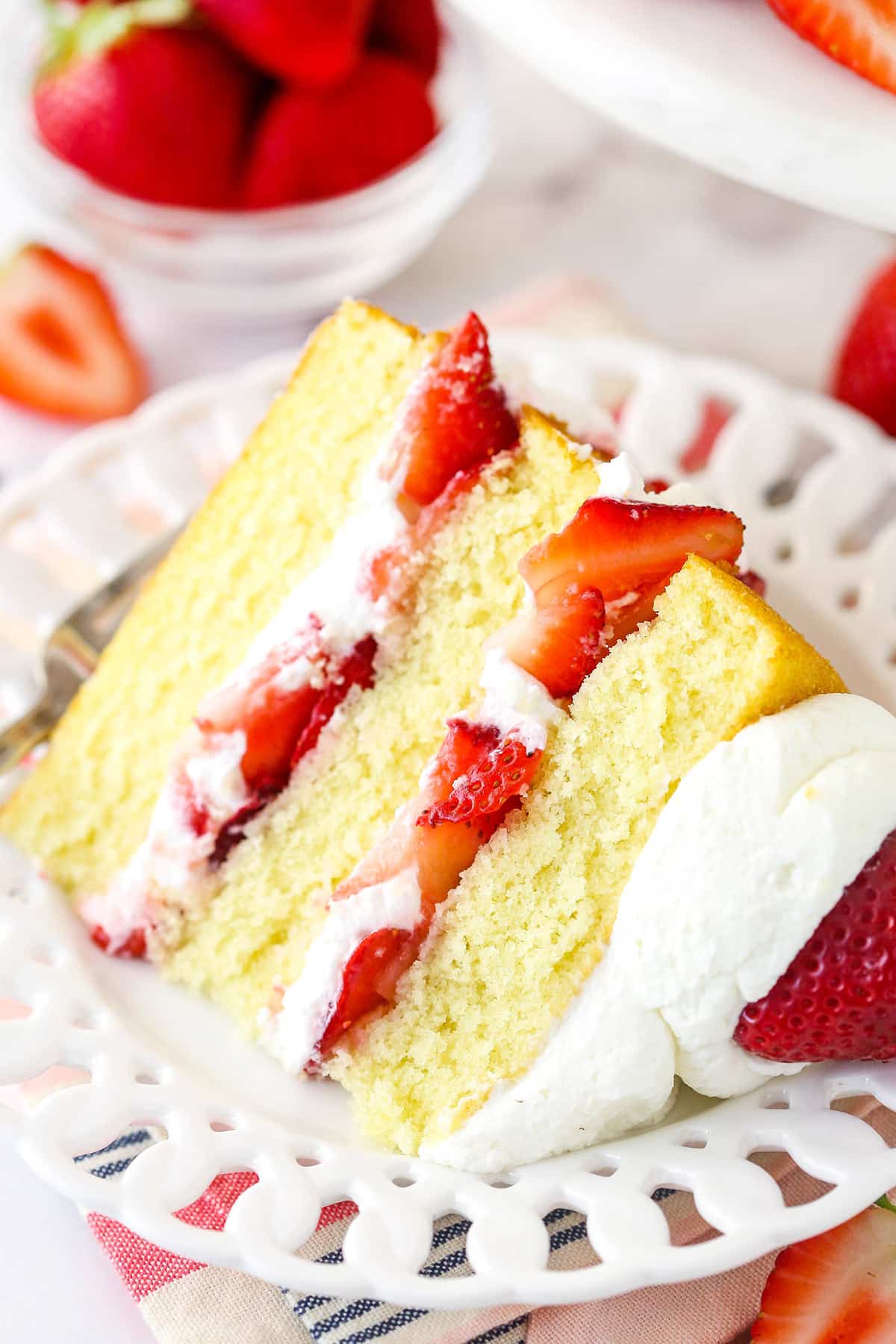 A slice of strawberry shortcake cake on a plate with a fork near fresh strawberries.