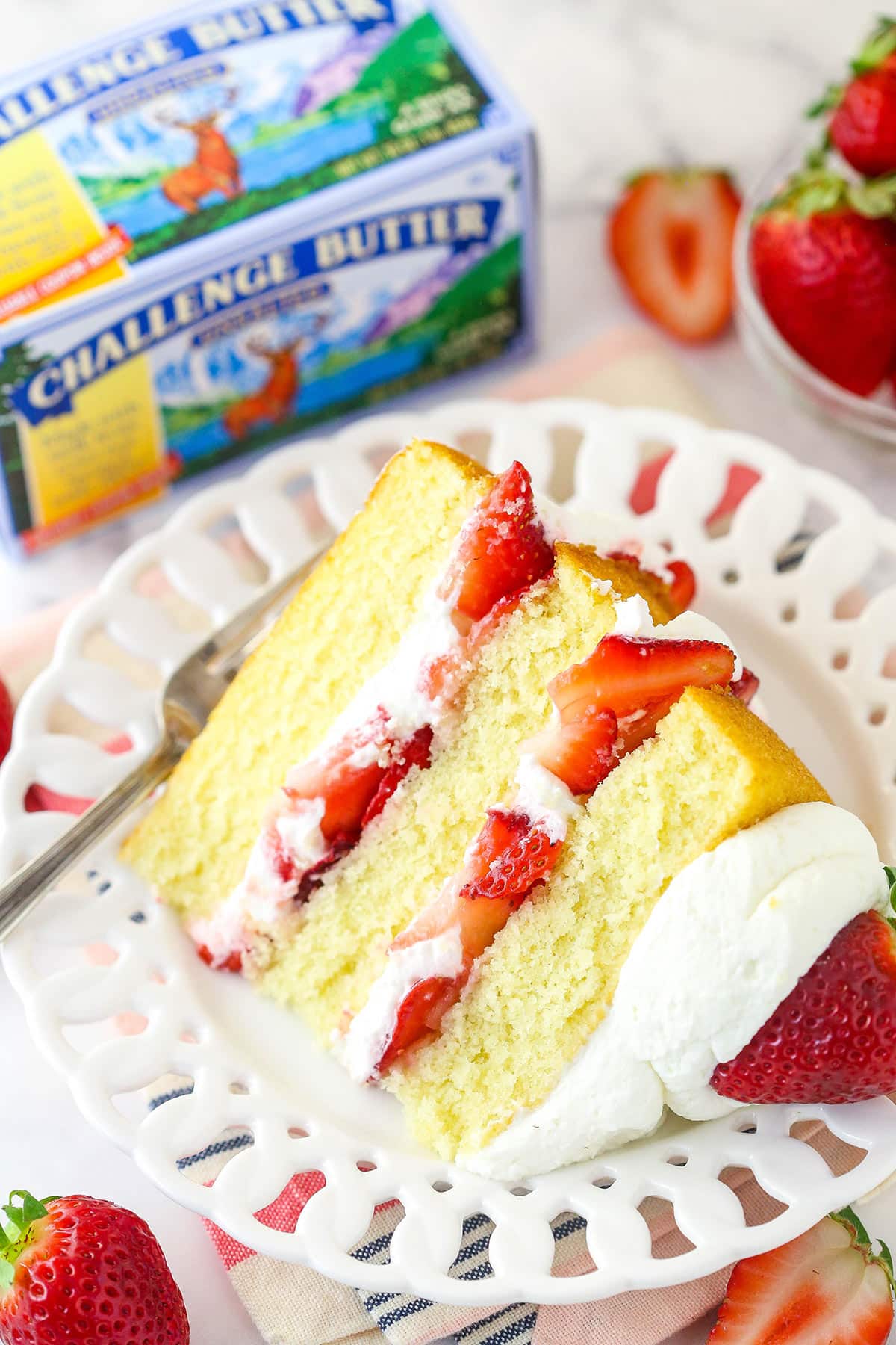 A slice of strawberry shortcake cake on a plate with a fork near a box of Challenge Butter.