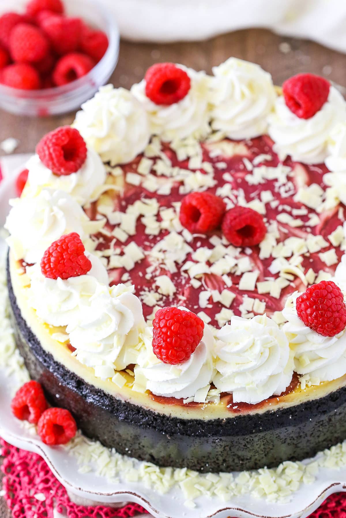 A full White Chocolate Raspberry Cheesecake with white swirls and raspberries on top, served on a white platter