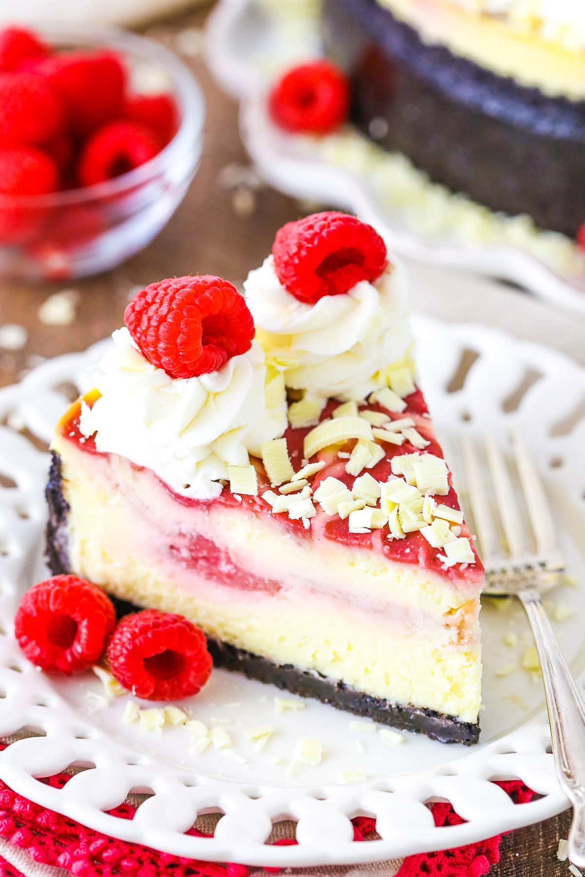 This White Chocolate Raspberry Cheesecake is a creamy white chocolate cheesecake swirled with raspberry filling, all set in a chocolate cookie crust! It's topped with whipped cream and white chocolate shavings for an amazingly tasty treat! A slice of White Chocolate Raspberry Cheesecake next to a fork on a white plate