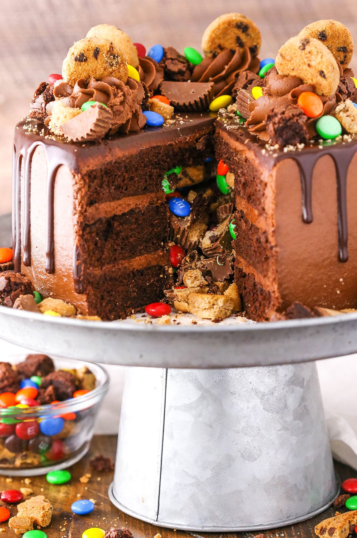 This Chocolate Piñata Cake is a moist chocolate cake frosted with classic chocolate buttercream and filled to the brim with awesome goodies - Reese's, M&Ms and chocolate chip cookies! Side view of a Chocolate Piñata Cake with a slice removed on a gray cake stand