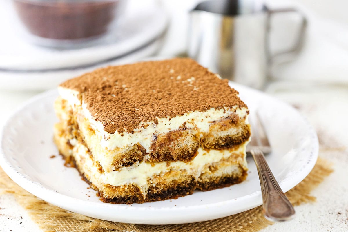 This easy tiramisu recipe is beyond decadent! Made with layers of espresso-soaked Ladyfinger cookies, fluffy mascarpone cream, and plenty of chocolate. A square serving of Easy Tiramisu next to a fork on a small white plate