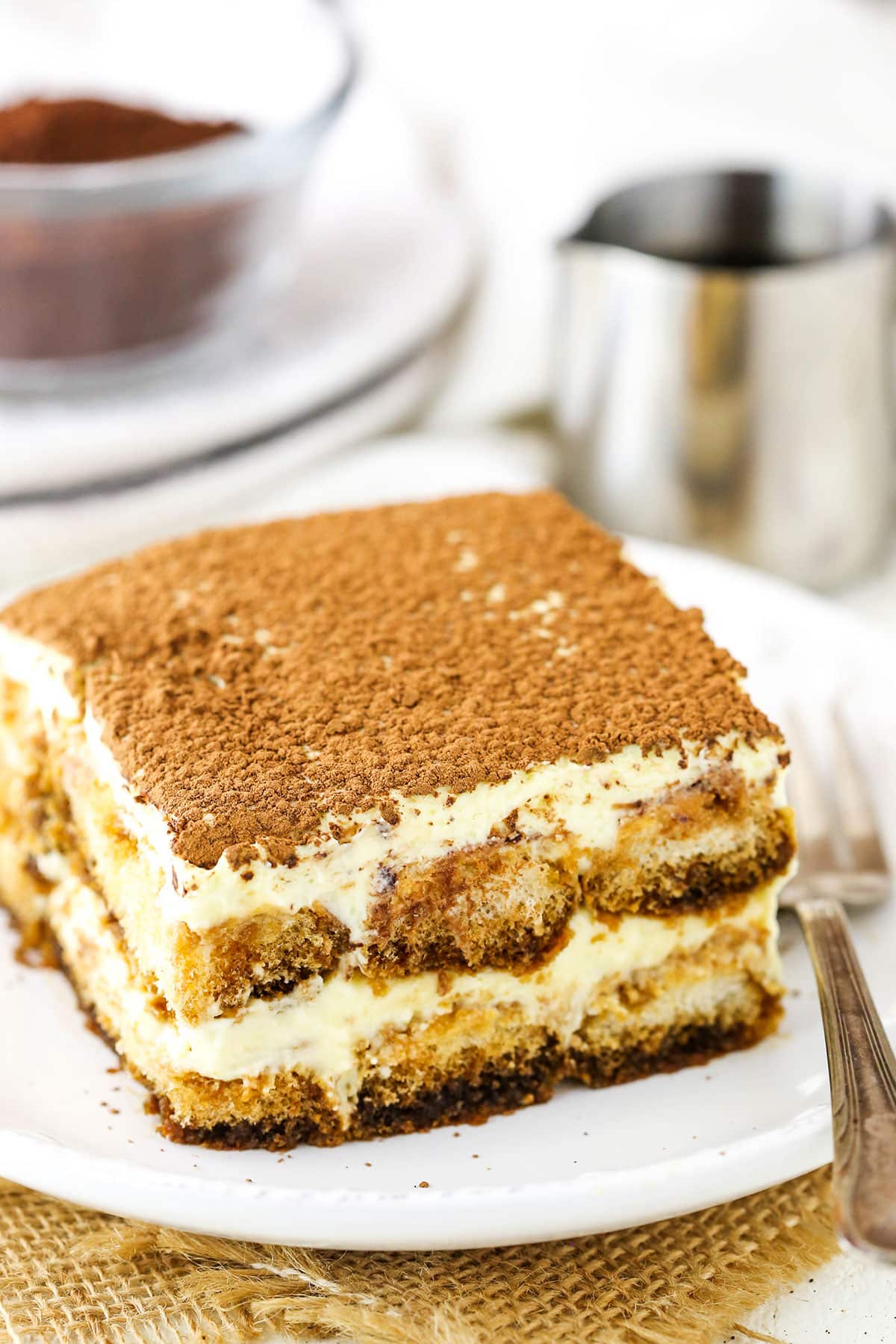 This easy tiramisu recipe is beyond decadent! Made with layers of espresso-soaked Ladyfinger cookies, fluffy mascarpone cream, and plenty of chocolate. A square serving of Easy Tiramisu next to a fork on a small white plate