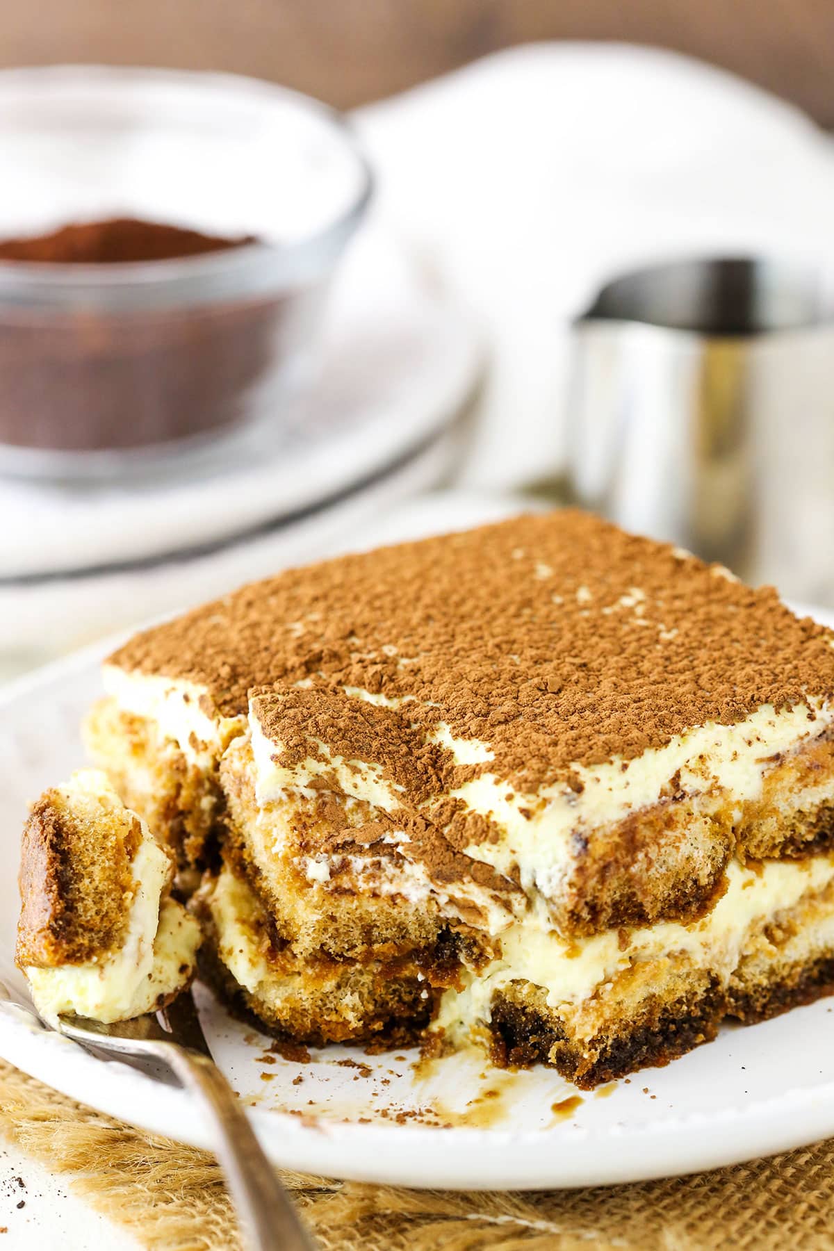 This easy tiramisu recipe is beyond decadent! Made with layers of espresso-soaked Ladyfinger cookies, fluffy mascarpone cream, and plenty of chocolate. A square serving of Easy Tiramisu with a bite taken out next to a fork on a small white plate