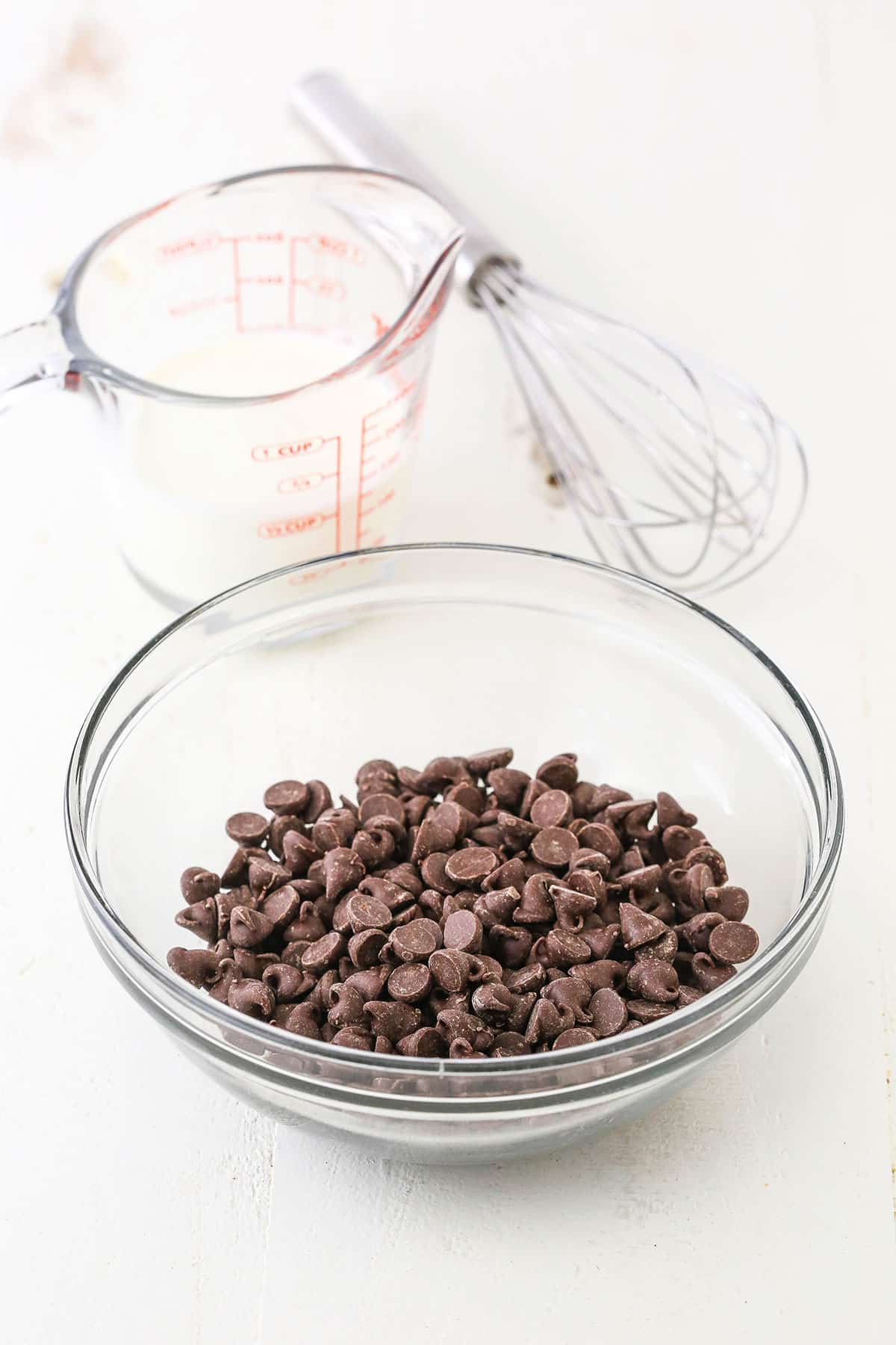 Chocolate chips in a clear glass bowl and heavy whipping cream in a clear glass measuring cup next to a whisk