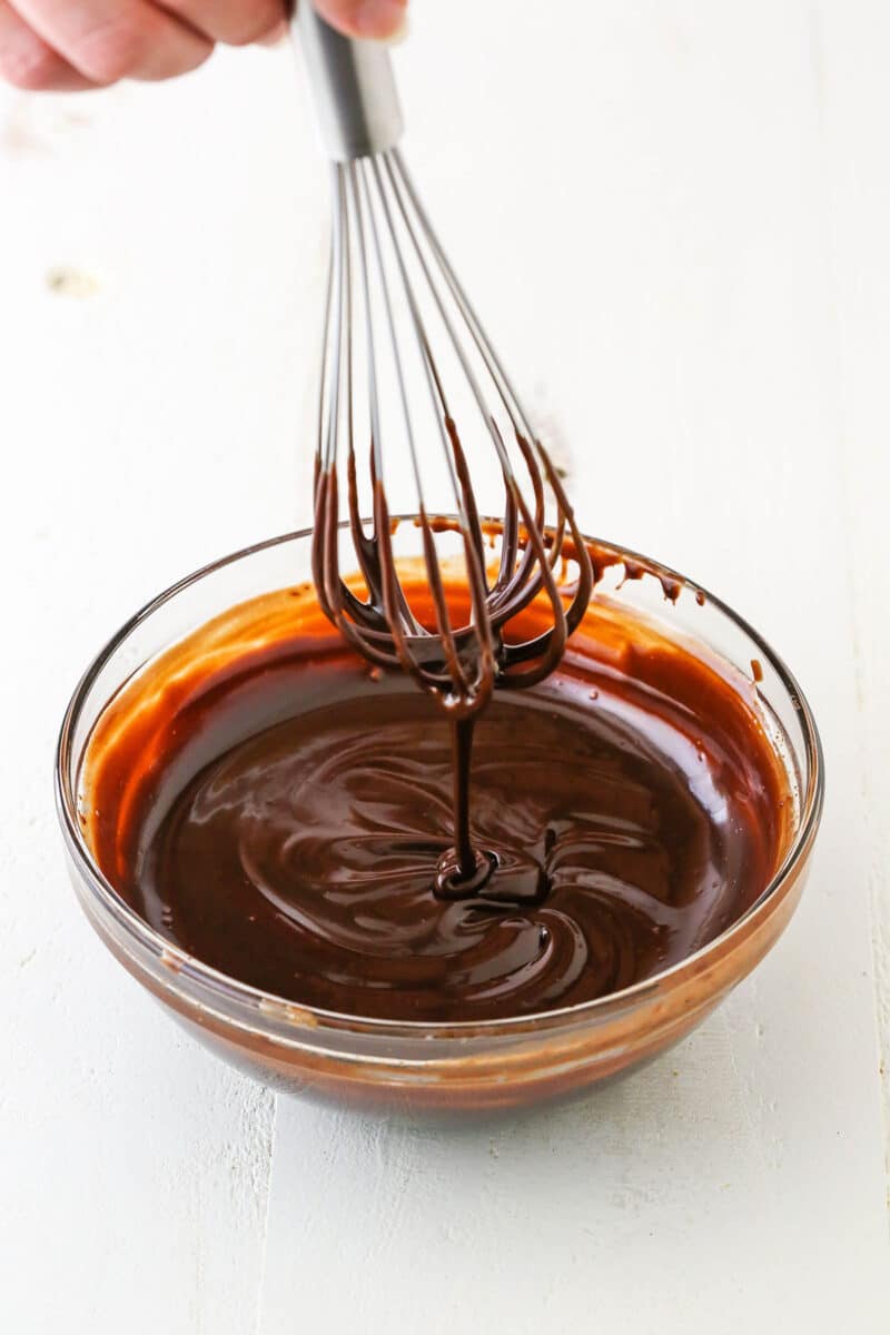 This Millionaire Cheesecake is next level! It's a tangy cheesecake doused in caramel and chocolate ganache atop a buttery shortbread crust. Dripping chocolate ganache off a whisk into a clear glass bowl