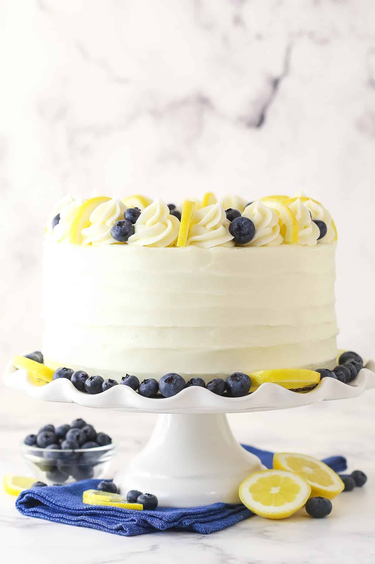 Lemon blueberry cake on a cake stand near fresh blueberries and lemon slices.
