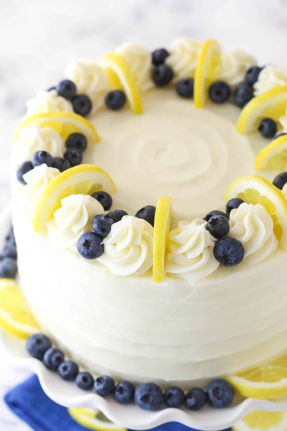 Overhead of lemon blueberry layer cake on a cake stand surrounded by fresh blueberries and lemon slices.