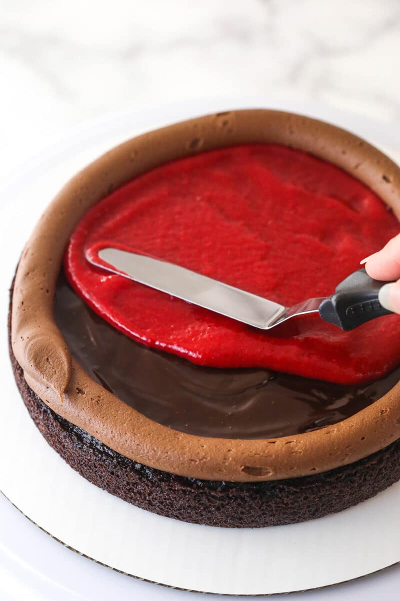 This chocolate raspberry cake is made of moist chocolate cake, smooth raspberry filling, dark chocolate frosting, and rich chocolate ganache. Spreading raspberry filling over chocolate ganache inside a dam of frosting on top of a layer of chocolate cake.