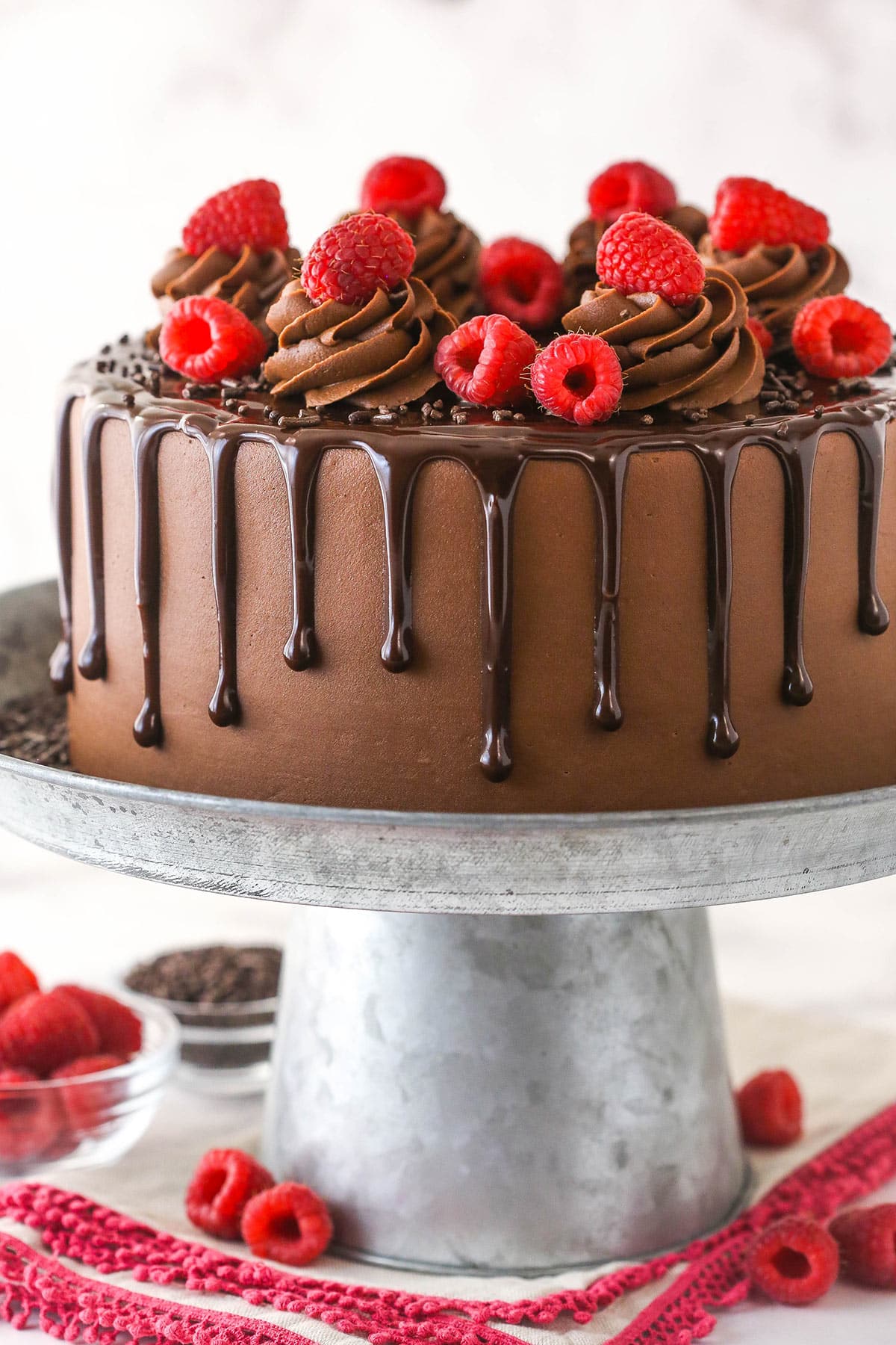 This chocolate raspberry cake is made of moist chocolate cake, smooth raspberry filling, dark chocolate frosting, and rich chocolate ganache. A whole raspberry chocolate take on a cake stand surrounded by fresh raspberries.