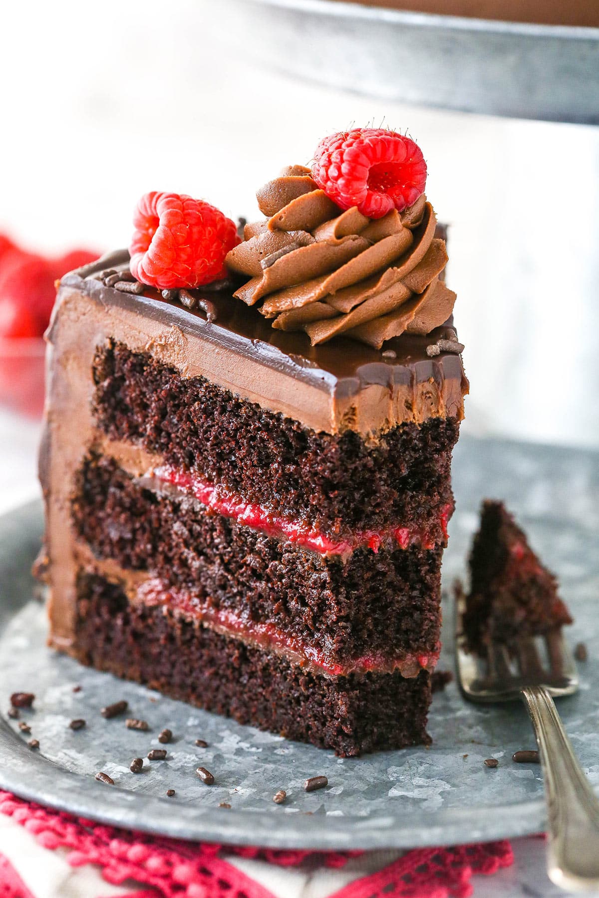 This chocolate raspberry cake is made of moist chocolate cake, smooth raspberry filling, dark chocolate frosting, and rich chocolate ganache. A slice of chocolate raspberry layer cake on a plate with a fork taking a bite out of it.