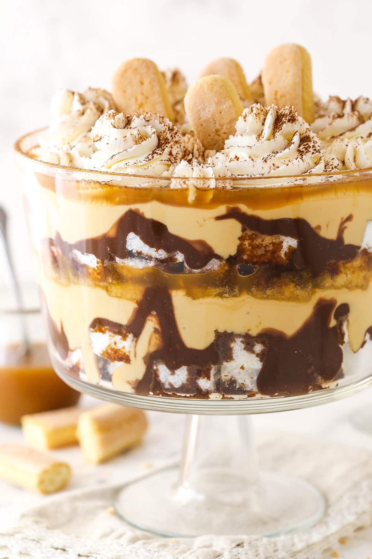 This quick and easy tiramisu trifle is made up of layers of ladyfingers, Nutella ganache, espresso, butterscotch pudding, & caramel sauce. Tiramisu trifle with lady fingers and a jar of caramel sauce in the background.
