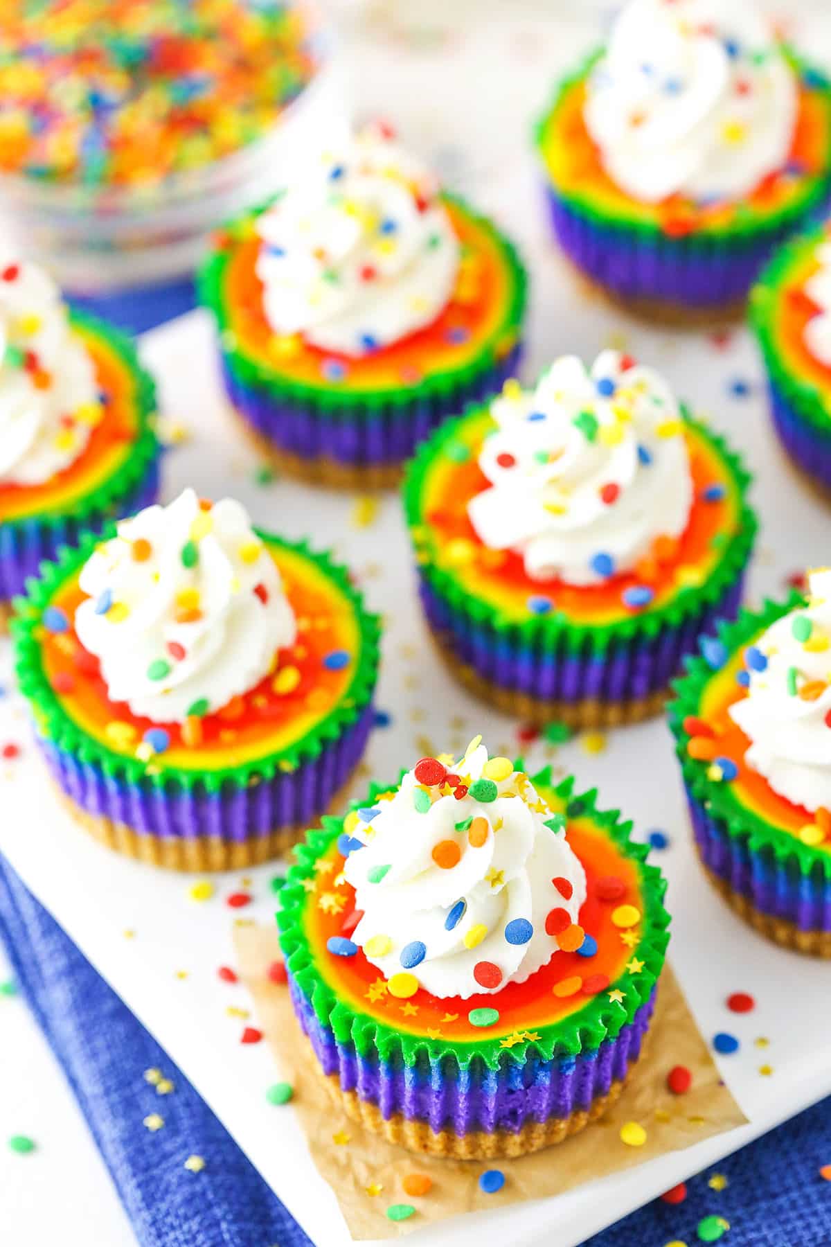 These Mini Rainbow Cheesecakes are so fun with colorful swirl layers! This fun cheesecake recipe is perfect for St. Patricks Day or a kid's party! Rainbow cake meets cheesecake! Overhead view of Mini Rainbow Cheesecakes on a white serving tray