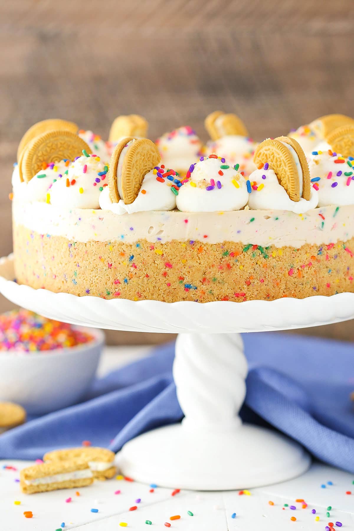 Side view of a full No-Bake Funfetti Oreo Birthday Cake Cheesecake on a white cake stand.