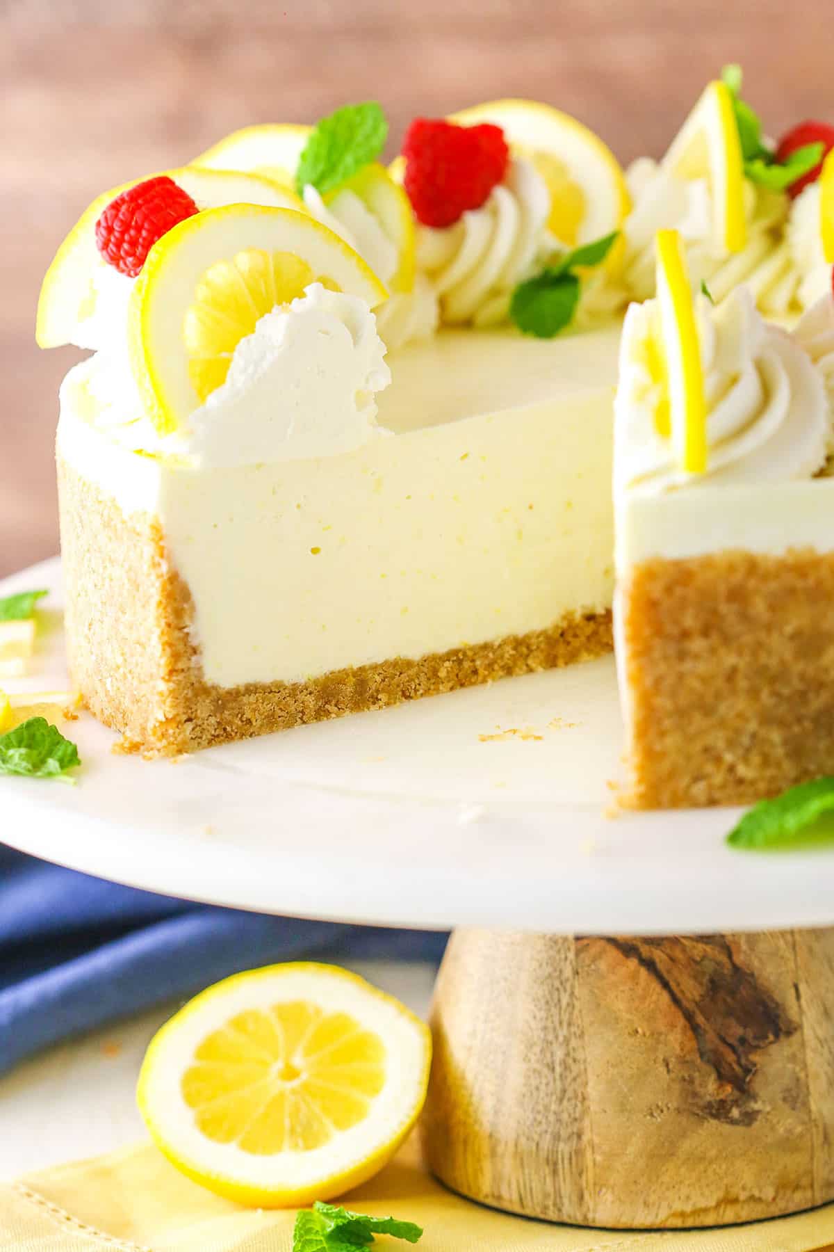 Side view of a No Bake Lemon Cheesecake with a slice removed on a white cake stand with a wooden base