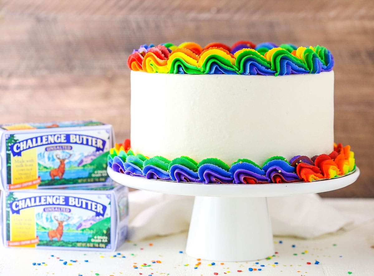 Side view of a full Rainbow Swirl Cake on a white cake stand next to two boxes of Challenge Butter