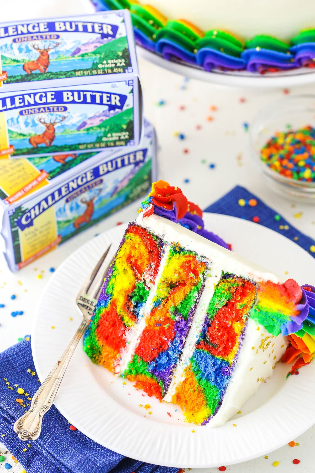 A slice of Rainbow Swirl Cake next to a fork on a white plate with Challenge Butter in the background