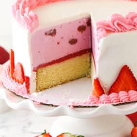Strawberry ice cream cake on a cake stand with a couple of slices taken out of it.