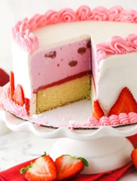 Strawberry ice cream cake on a cake stand with a couple of slices taken out of it.