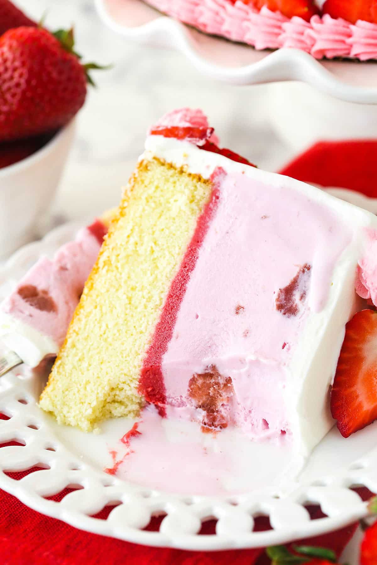 A slice of strawberry ice cream cake on a plate with a bite taken out of it.
