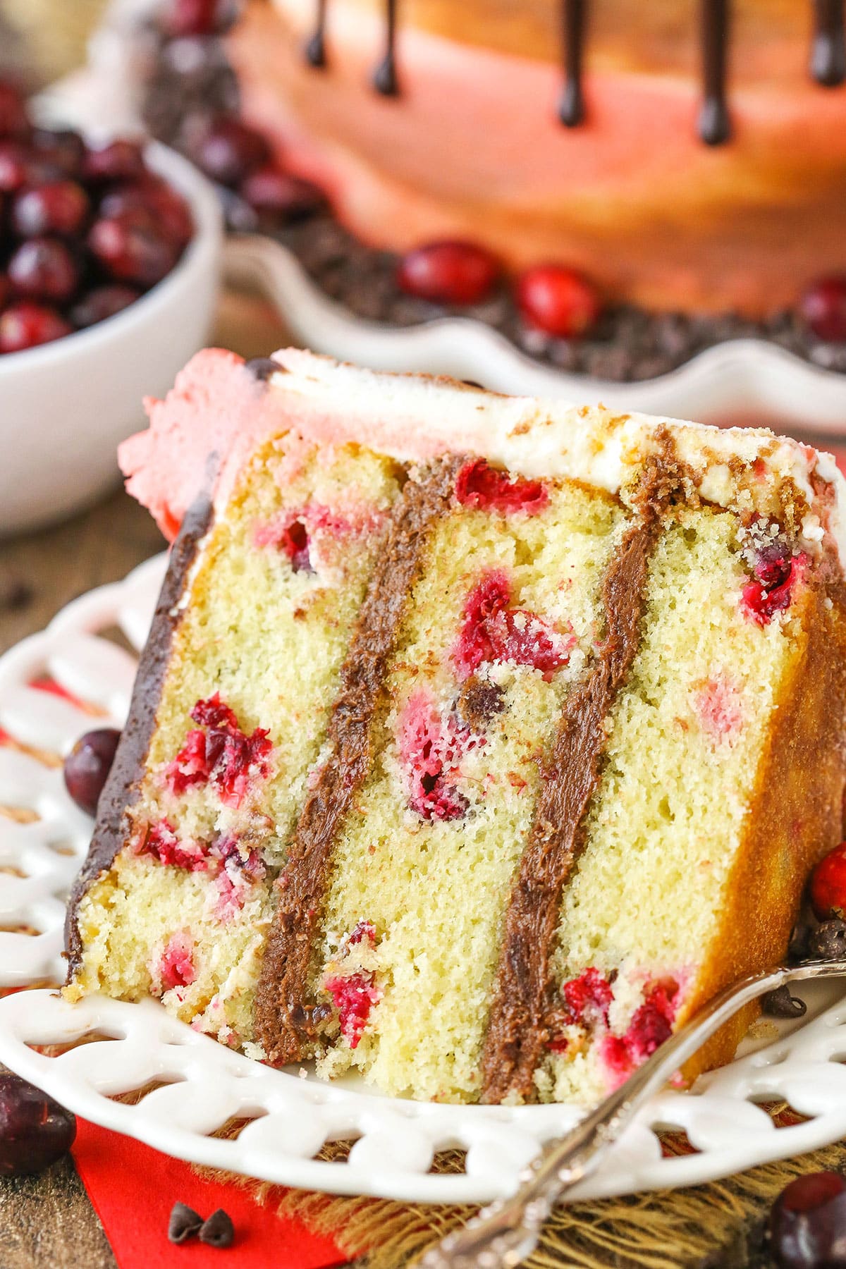 This Cranberry Fudge Layer Cake is made with a moist vanilla cake studded with fresh cranberries and filled with chocolate fudgy frosting! It's a moist, delicious cake and great for the holidays! A slice of Cranberry Fudge Layer Cake next to a fork on a white plate
