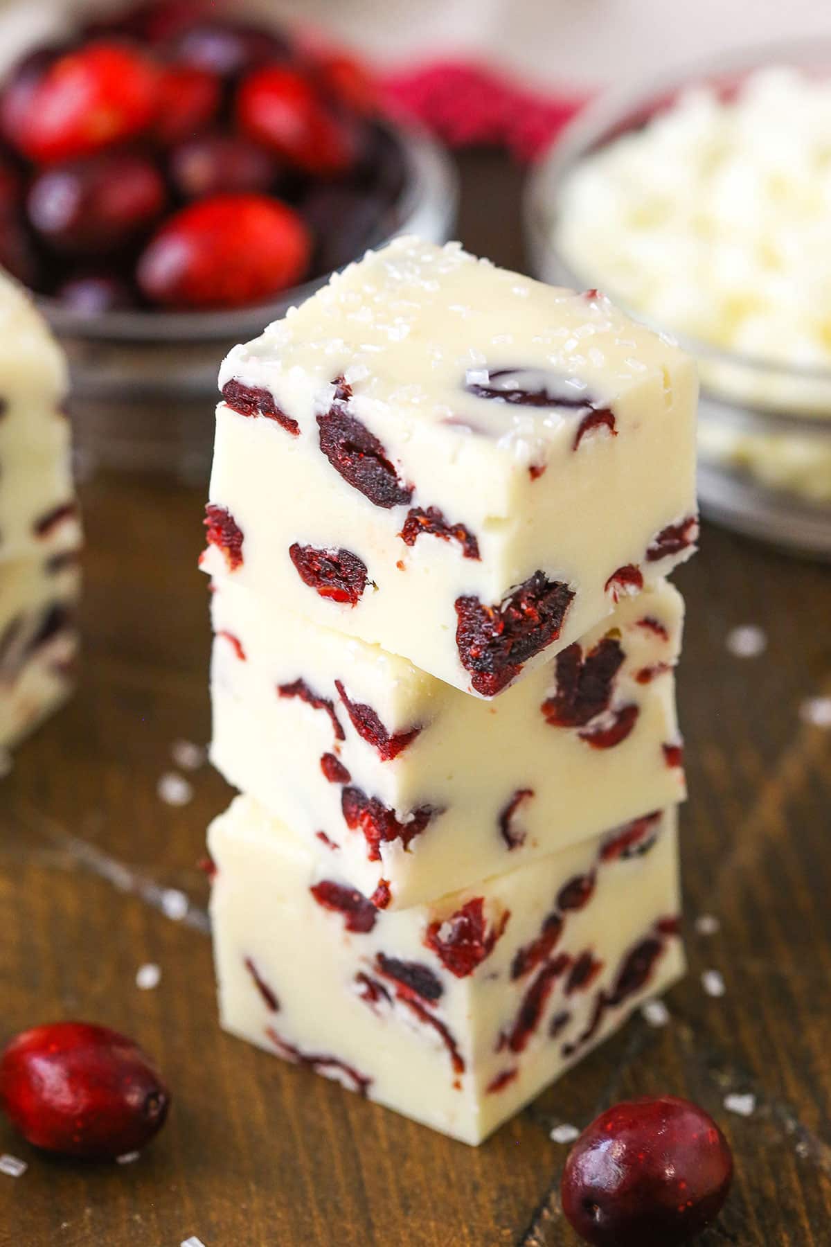 This White Chocolate Cranberry Fudge is made with sweetened condensed milk and dried cranberries! With beautiful Christmas colors, this super easy and quick dessert is a great treat for the busy holiday season! Three White Chocolate Cranberry Fudge cubes stacked on a wooden table