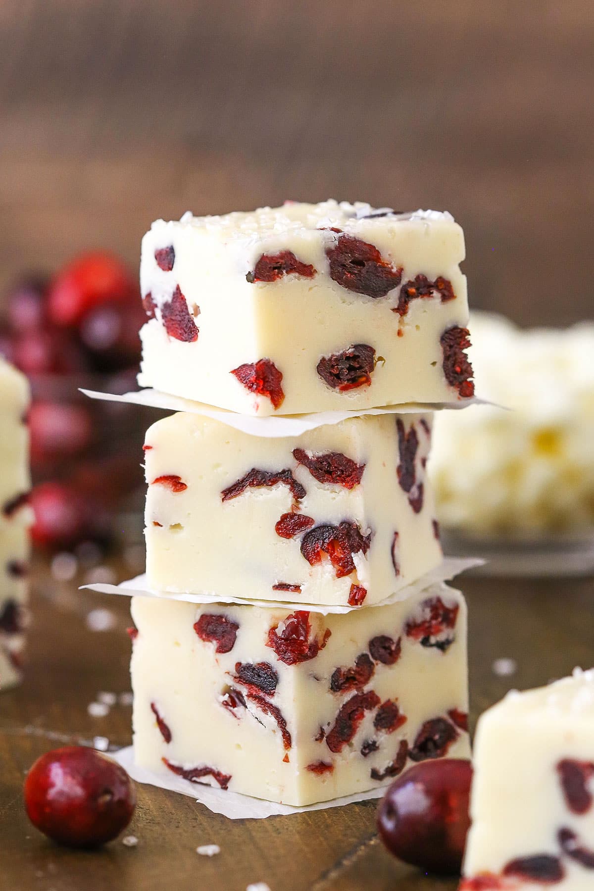 This White Chocolate Cranberry Fudge is made with sweetened condensed milk and dried cranberries! With beautiful Christmas colors, this super easy and quick dessert is a great treat for the busy holiday season! Three White Chocolate Cranberry Fudge cubes stacked on a wooden table