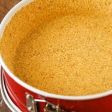 Graham cracker crust in a red springform pan on a wooden table