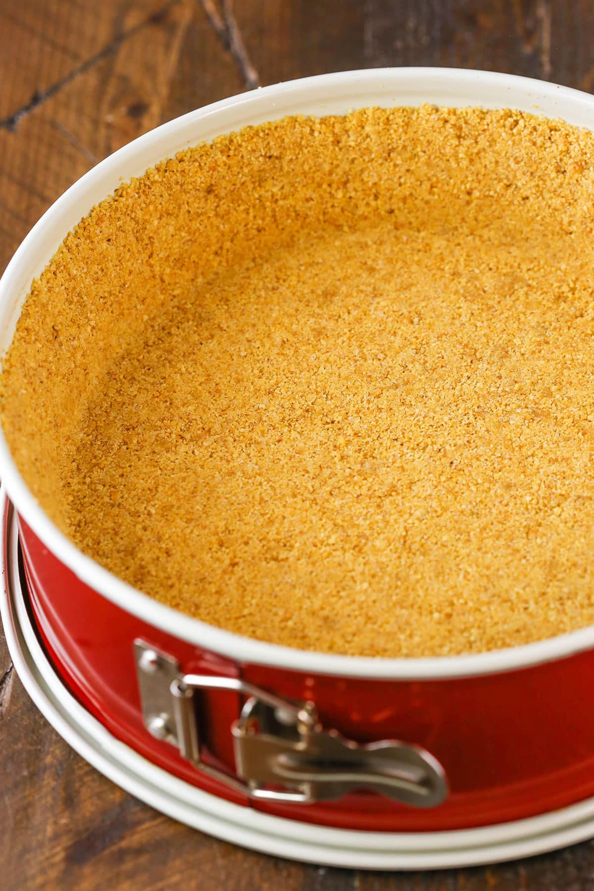 This Graham Cracker Crust recipe is easy to make and delicious! Made from scratch with only four ingredients, it won't fall apart and is perfect for baked pies, no bake pies and cheesecakes! Easy Graham Cracker Crust in a red springform pan on a wooden table