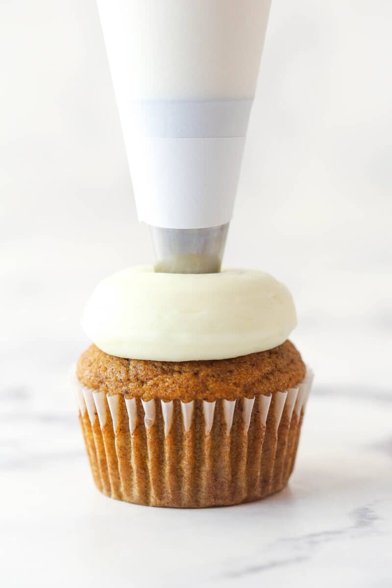 Piping cream cheese frosting onto a pumpkin cupcake.