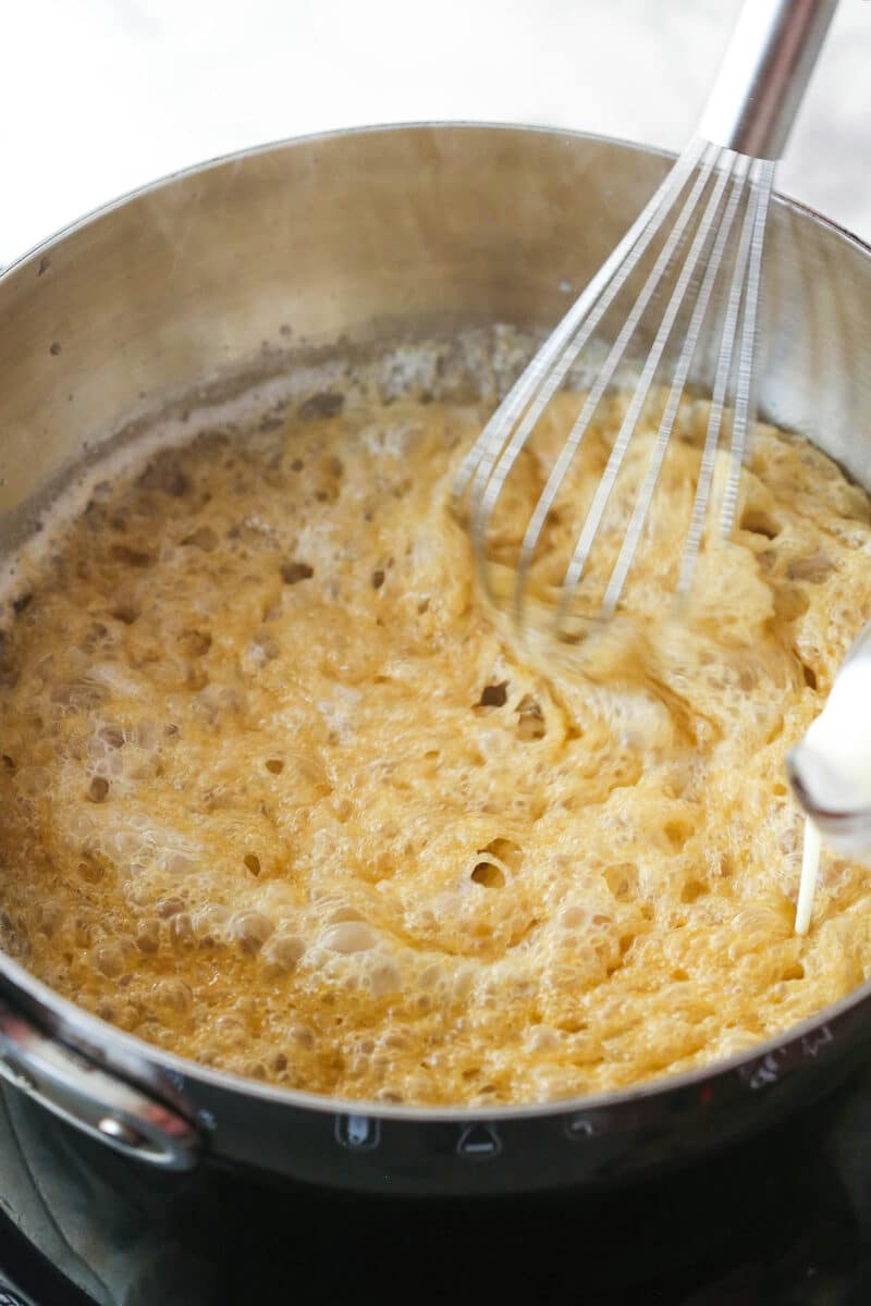 Pouring cream into a metal pot containing the sugar mixture and a whisk.