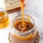 Bourbon Caramel Sauce dripping from a spoon into a jar filled with sauce with bourbon in the background.
