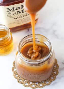 Bourbon Caramel Sauce dripping from a spoon into a jar filled with sauce with bourbon in the background.