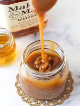 Bourbon Caramel Sauce dripping from a spoon into a jar filled with sauce with bourbon in the background.