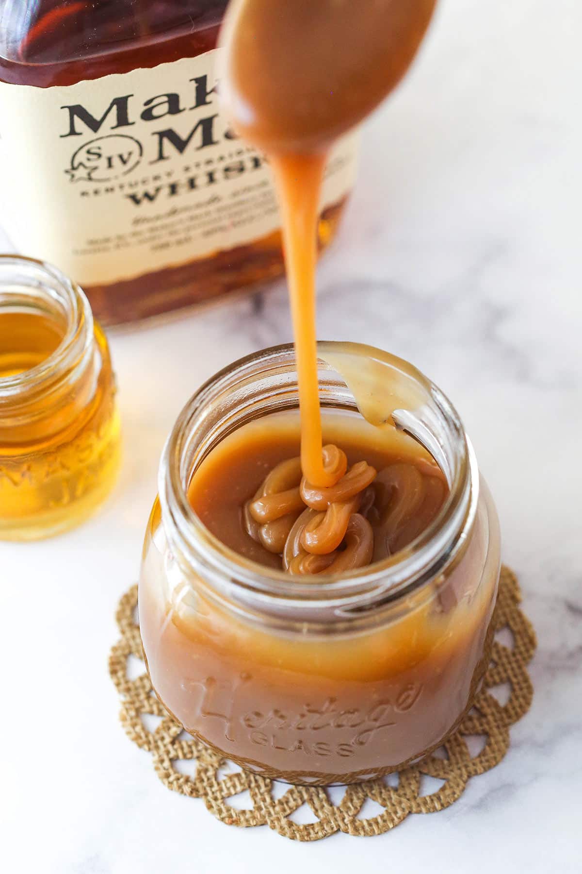 Bourbon Caramel Sauce dripping from a spoon into a jar filled with sauce with bourbon in the background.