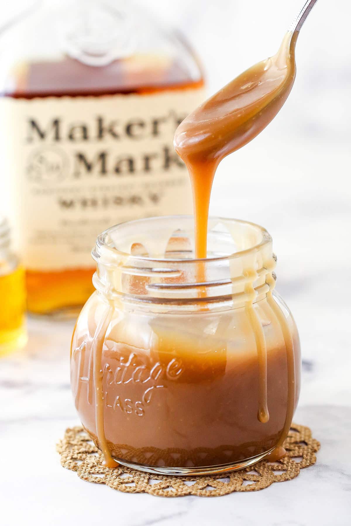 Side view of Bourbon Caramel Sauce dripping from a spoon into a jar filled with sauce with bourbon in the background.