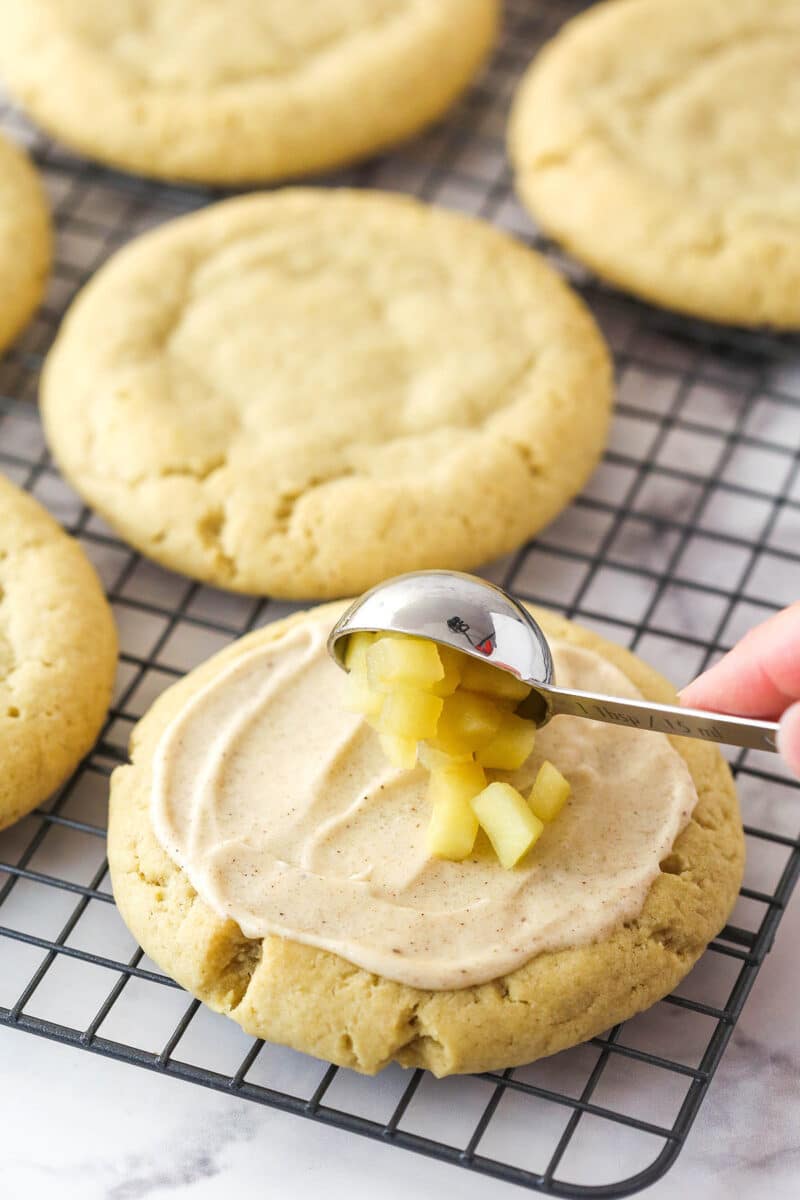 A scoop of apples being placed in the center of a frosted Copycat Crumbl Apple Crumb Cake Cookie.