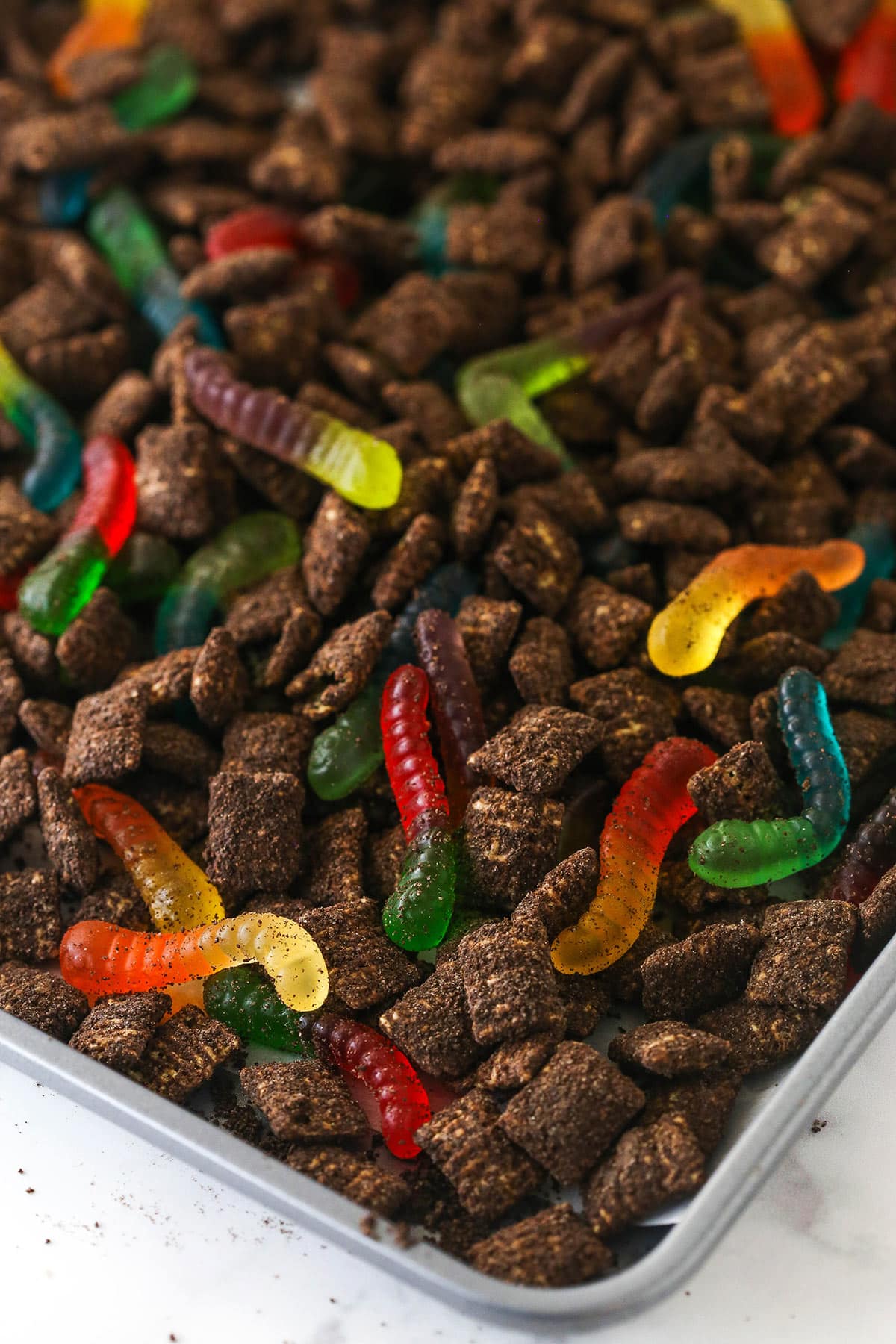 Chocolate covered Chex cereal and gummy worms laid out on a parchment paper-lined baking sheet for Dirt and Worms Puppy Chow.